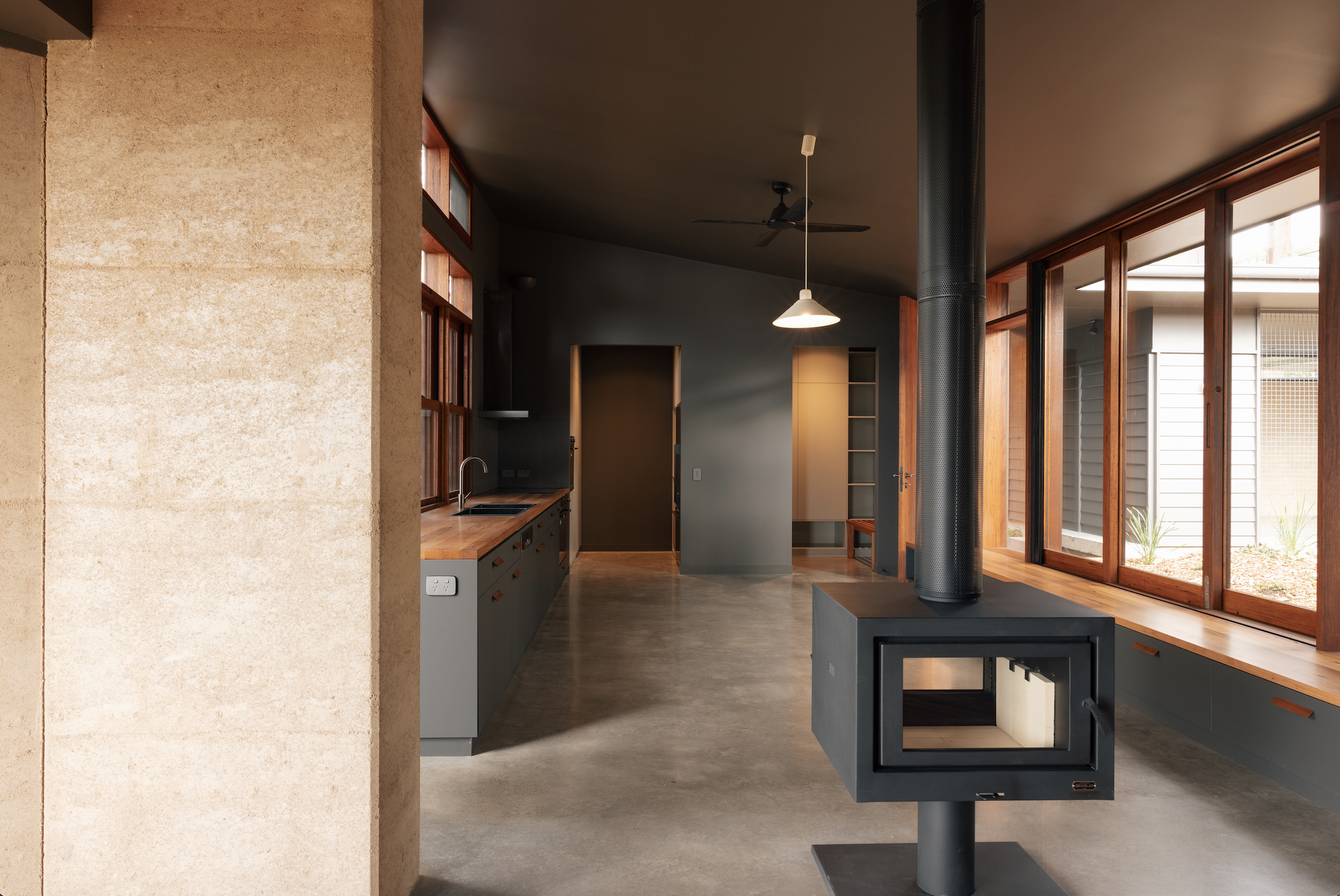 Open-plan living and kitchen — freestanding fireplace, rammed earth column, polished concrete and timber windows