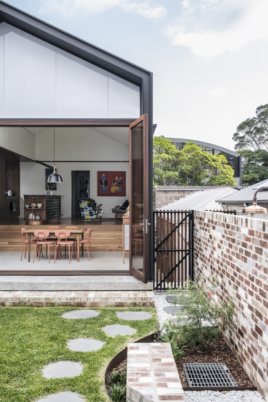 Garden view — bifold doors open to dining and living, stepping stones across lawn, recycled brick wall